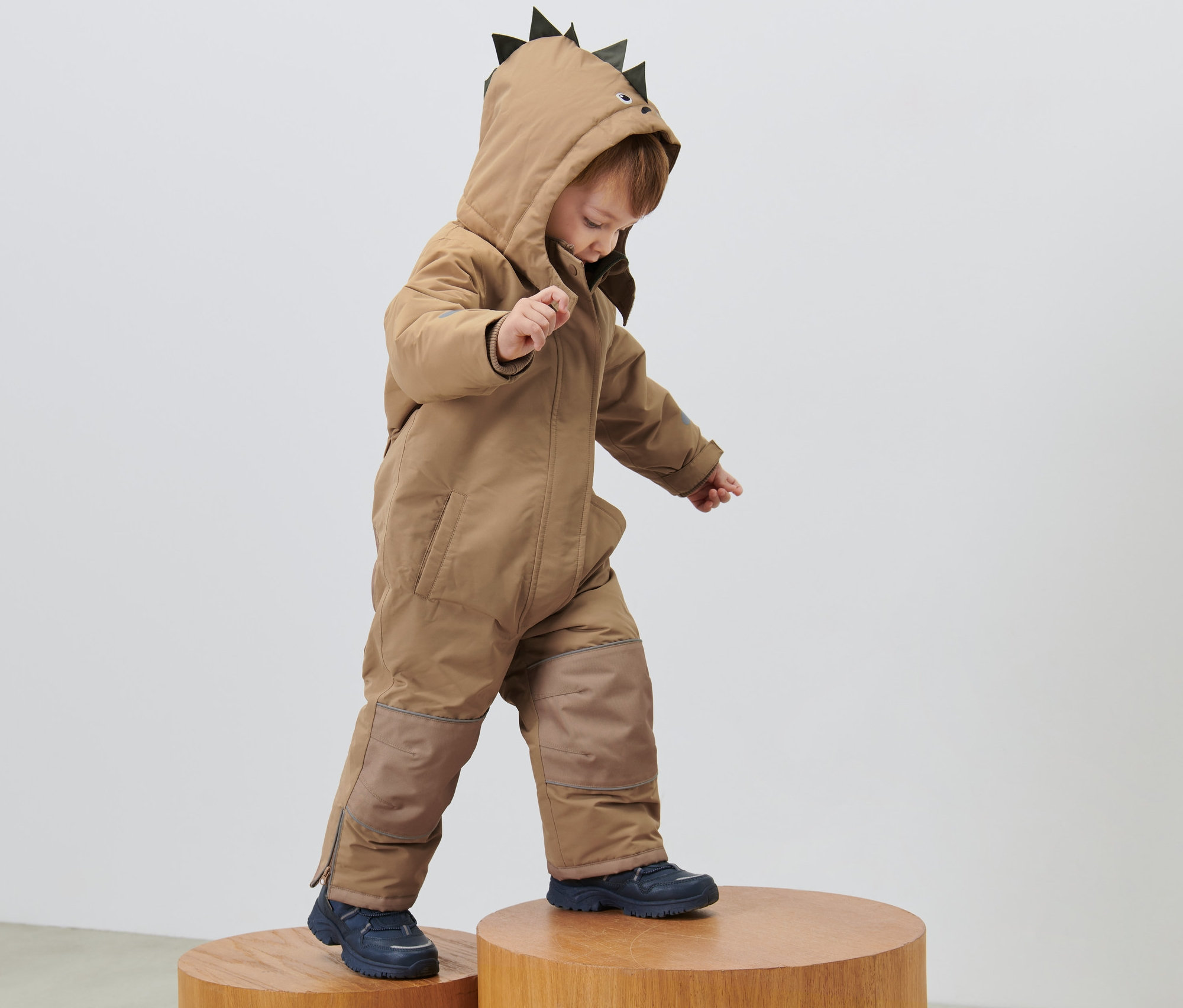 Çocuk, başlığında dikenleri olan çocuk kar tulumuyla ahşap taburelerin üzerinde duruyor.