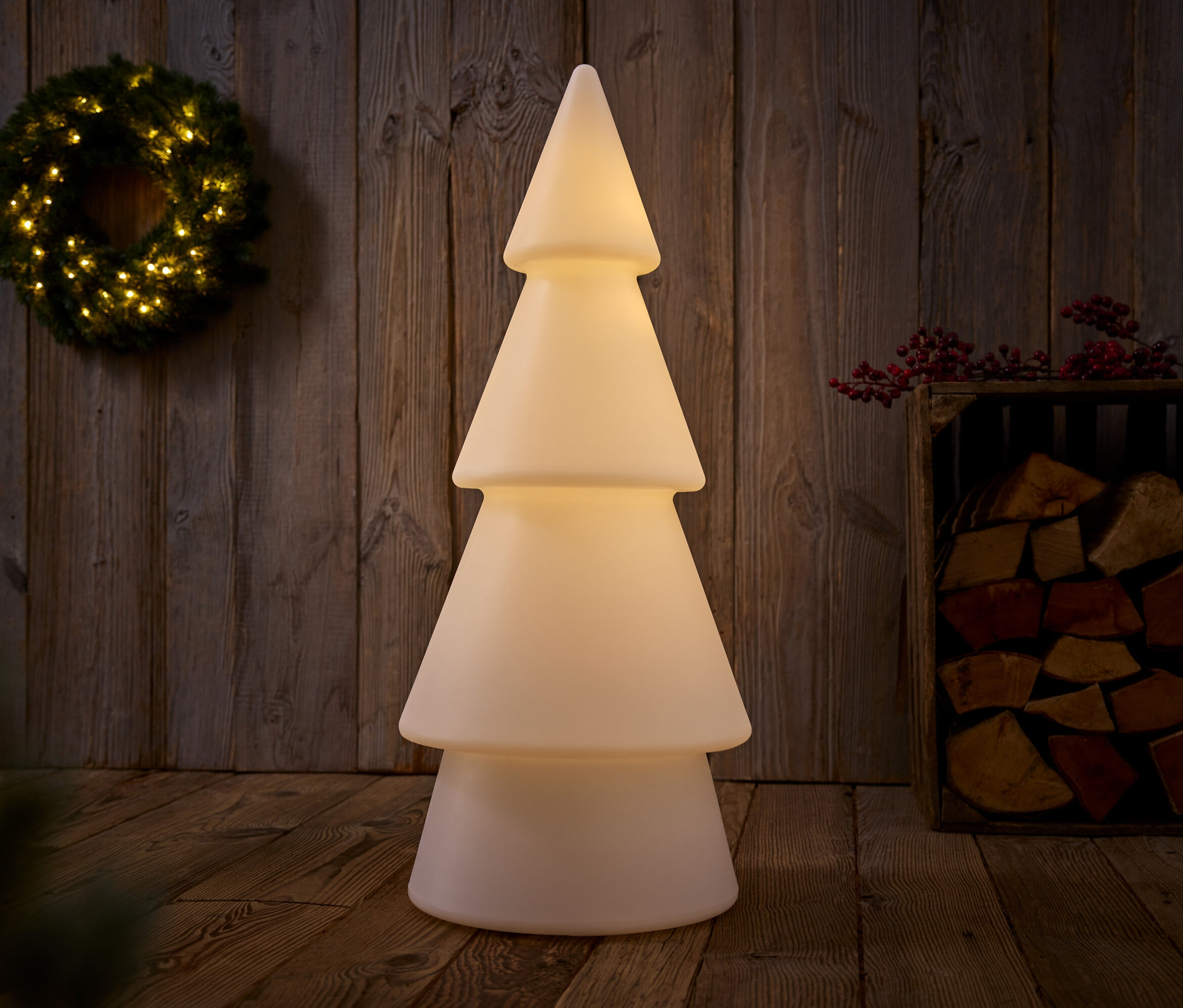 Ahşap bir sandık içinde odunların ve duvarda asılı bir çelenkin yanında duran, aydınlatılmış dekoratif bir Noel ağacının dikey çekimi.