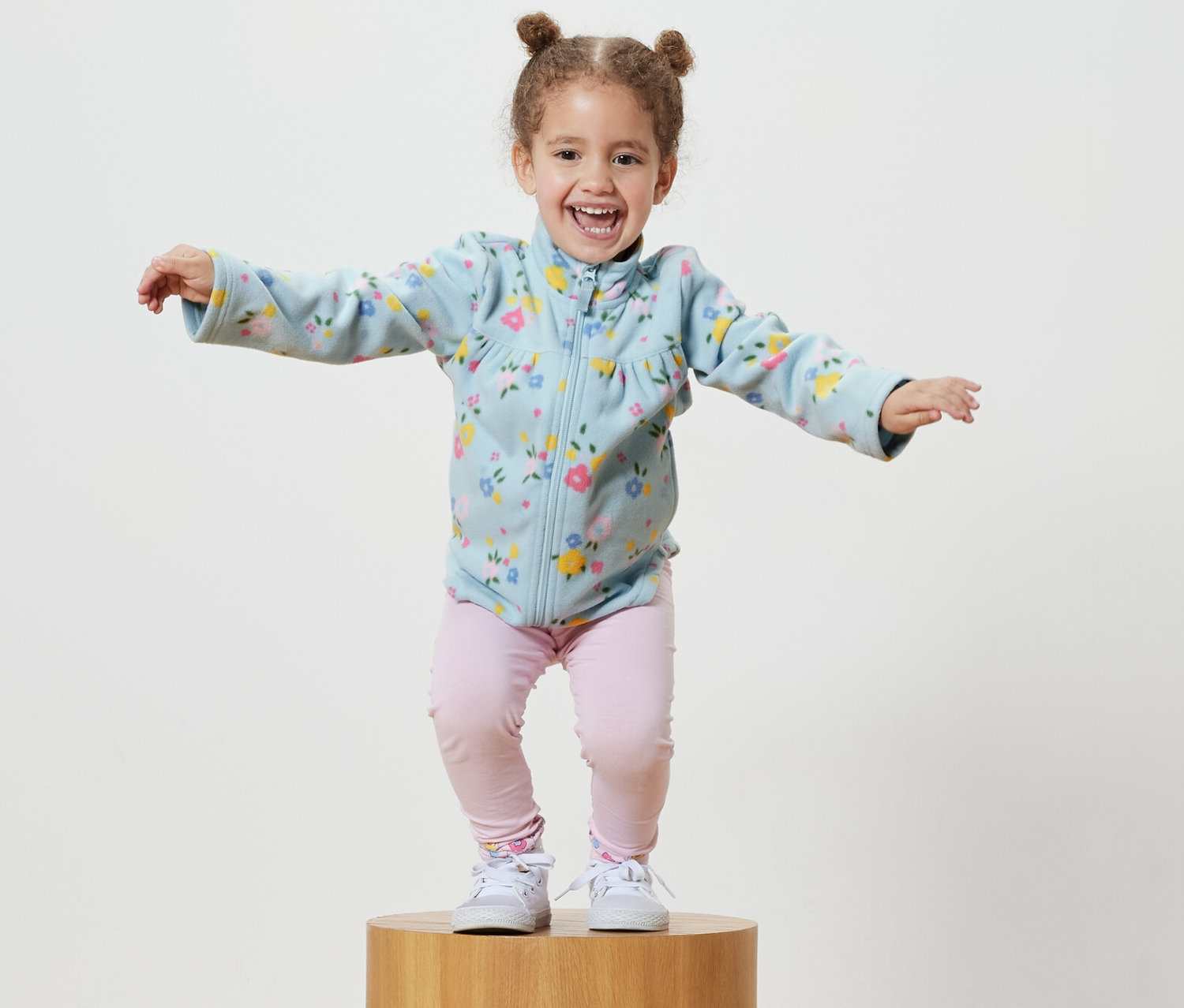 Gülümseyen küçük bir kız tahta bir kaide üzerinde duruyor ve açık mavi bir polar mont giyiyor.