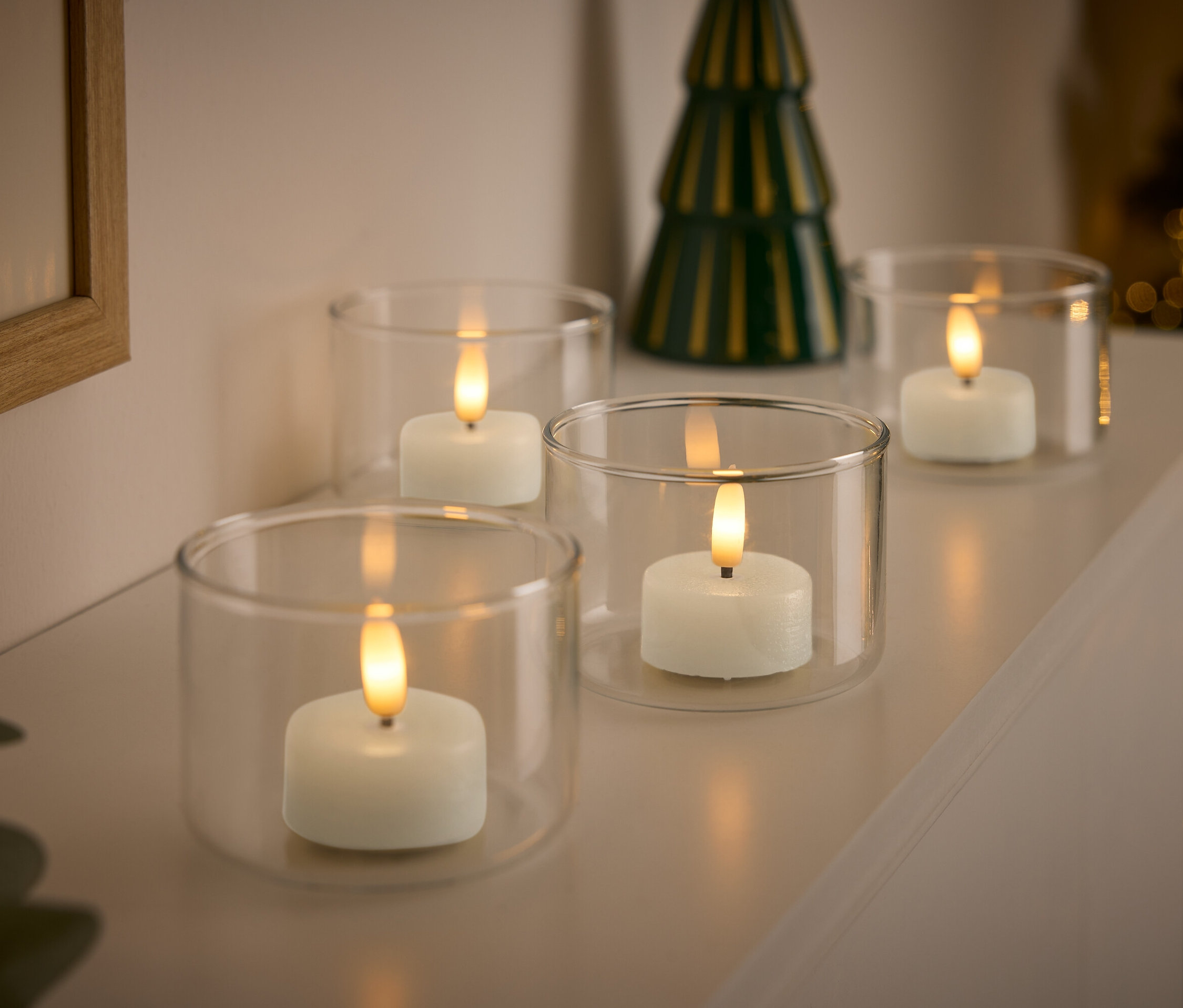 Arka planda küçük yeşil bir Noel ağacıyla birlikte şeffaf cam mumluklar içinde dört beyaz mum.