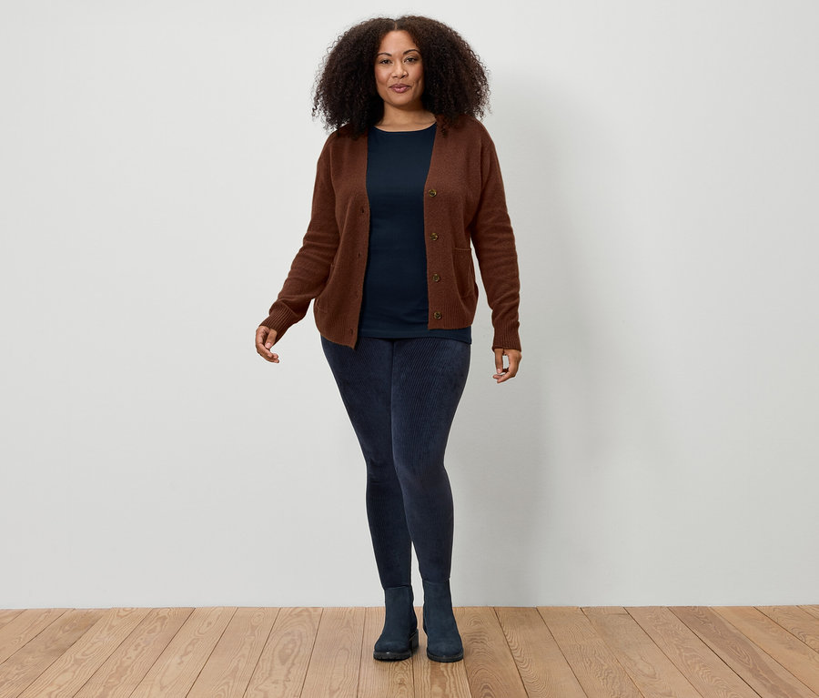 Kahverengi kıvırcık saçlı bir kadın, beyaz bir duvarın önünde duruyor ve kahverengi bir hırka, lacivert bir tişört, lacivert tayt ve lacivert botlar giyiyor.