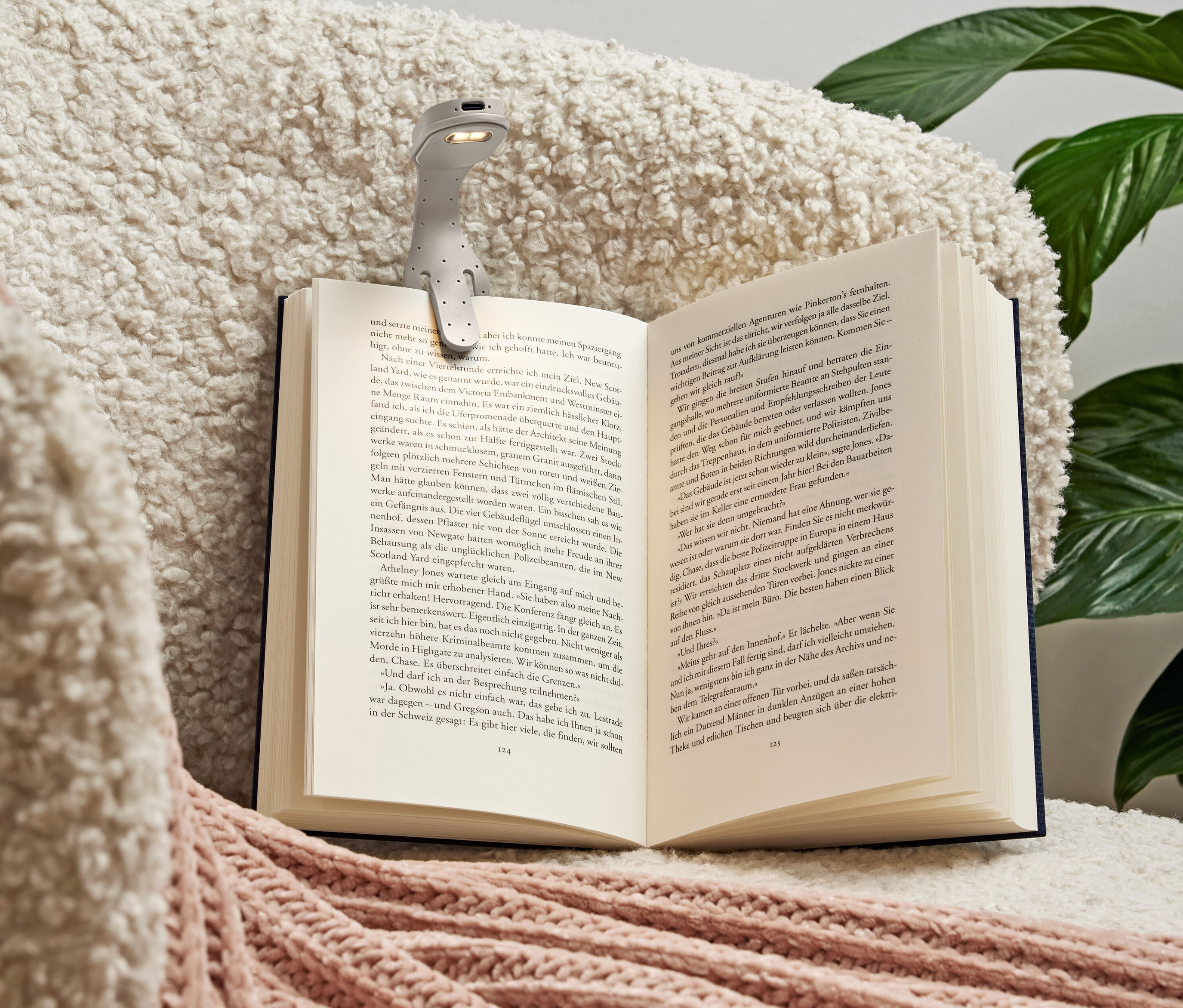 Gri bir okuma lambası, pembe detaylara sahip yumuşak, krem rengi bir battaniyenin üzerinde açık bir kitaba tutturulmuş.