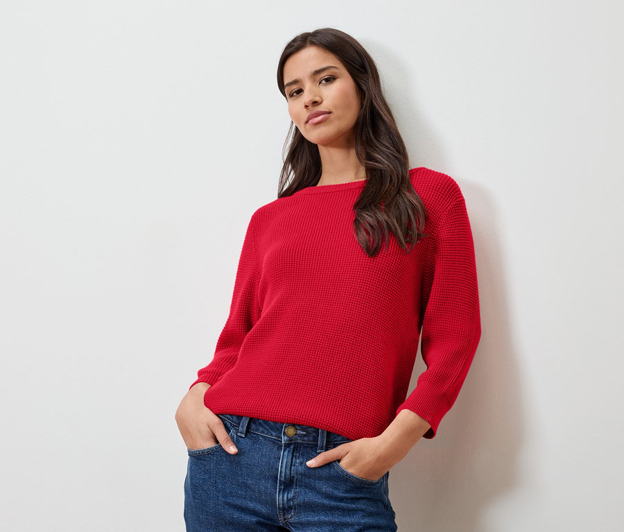 Beyaz bir duvarın önünde poz veren kırmızı ince triko kazak ve kot pantolonlu kadın.