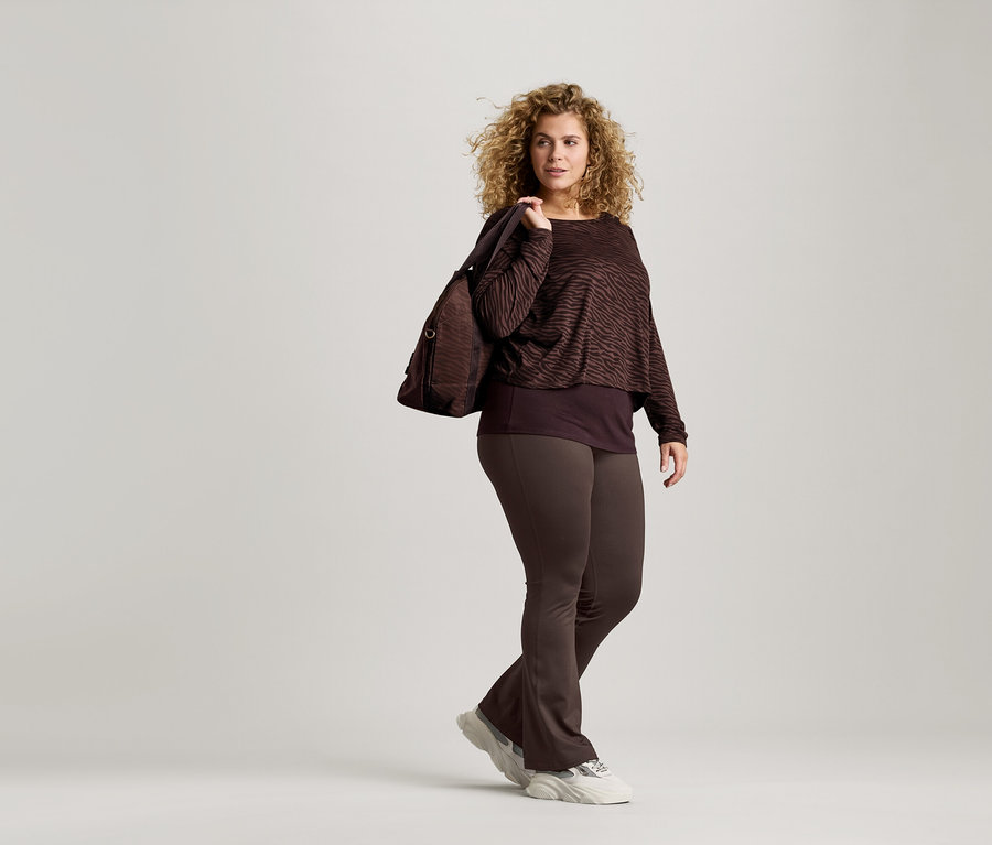 Kıvırcık saçlı kadın, zebra desenli kahverengi bir bluz, kahverengi tayt ve spor ayakkabı giyiyor. Omuzunda kahverengi bir çanta var.