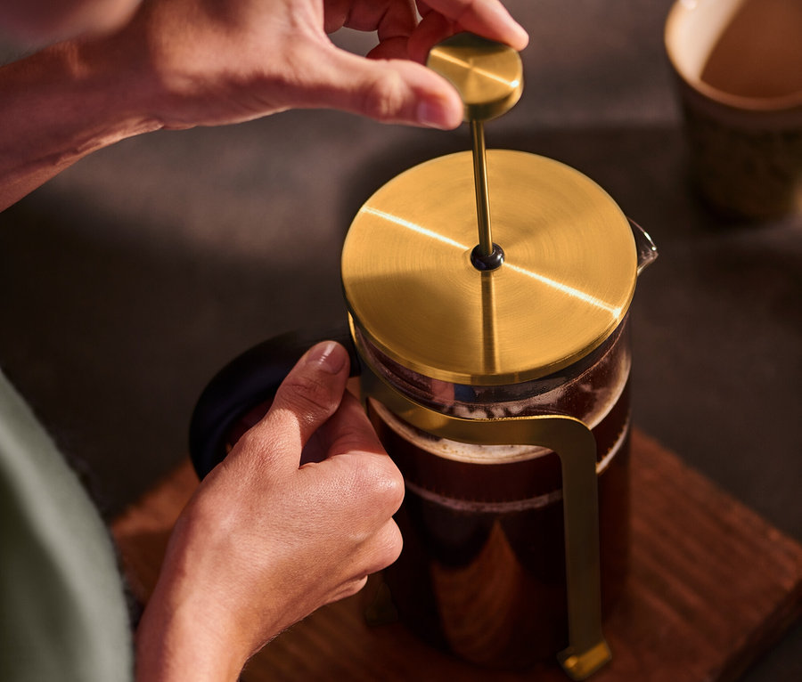 Bir kişi, ahşap bir altlık üzerinde altın rengi 800 ml'lik 6 fincanlık bir French press'i aşağı doğru itiyor.