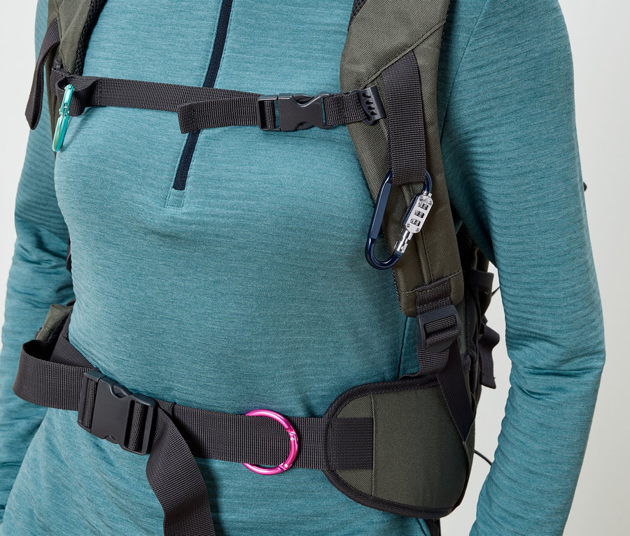 Bir kadın, üzerinde siyah kayışları ve tokaları olan zeytin yeşili bir sırt çantası takıyor; üzerinde şifreli kilitli mavi bir karabina ve turkuaz bir karabina takılı.