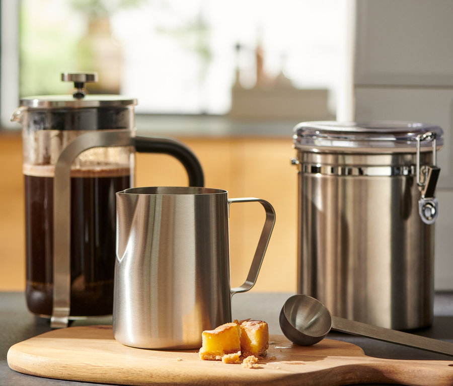 Bir bardak kahve, ahşap bir kesme tahtası üzerinde bir french press ve küçük bir paslanmaz çelik sütlüğün yanında duruyor.