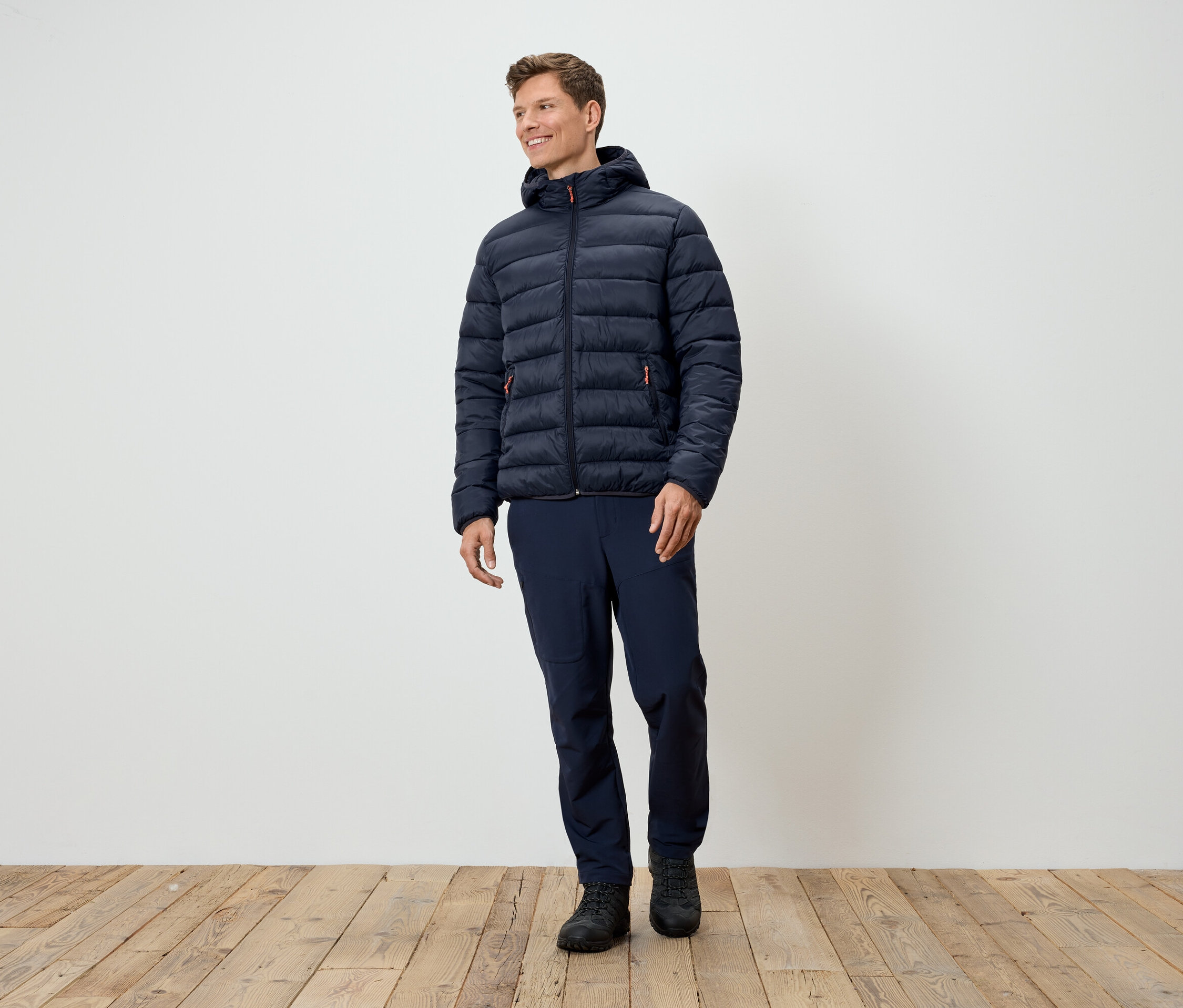 Bir adam ayakta duruyor ve lacivert kapitone ceket, lacivert pantolon ve siyah ayakkabılar giyiyor.