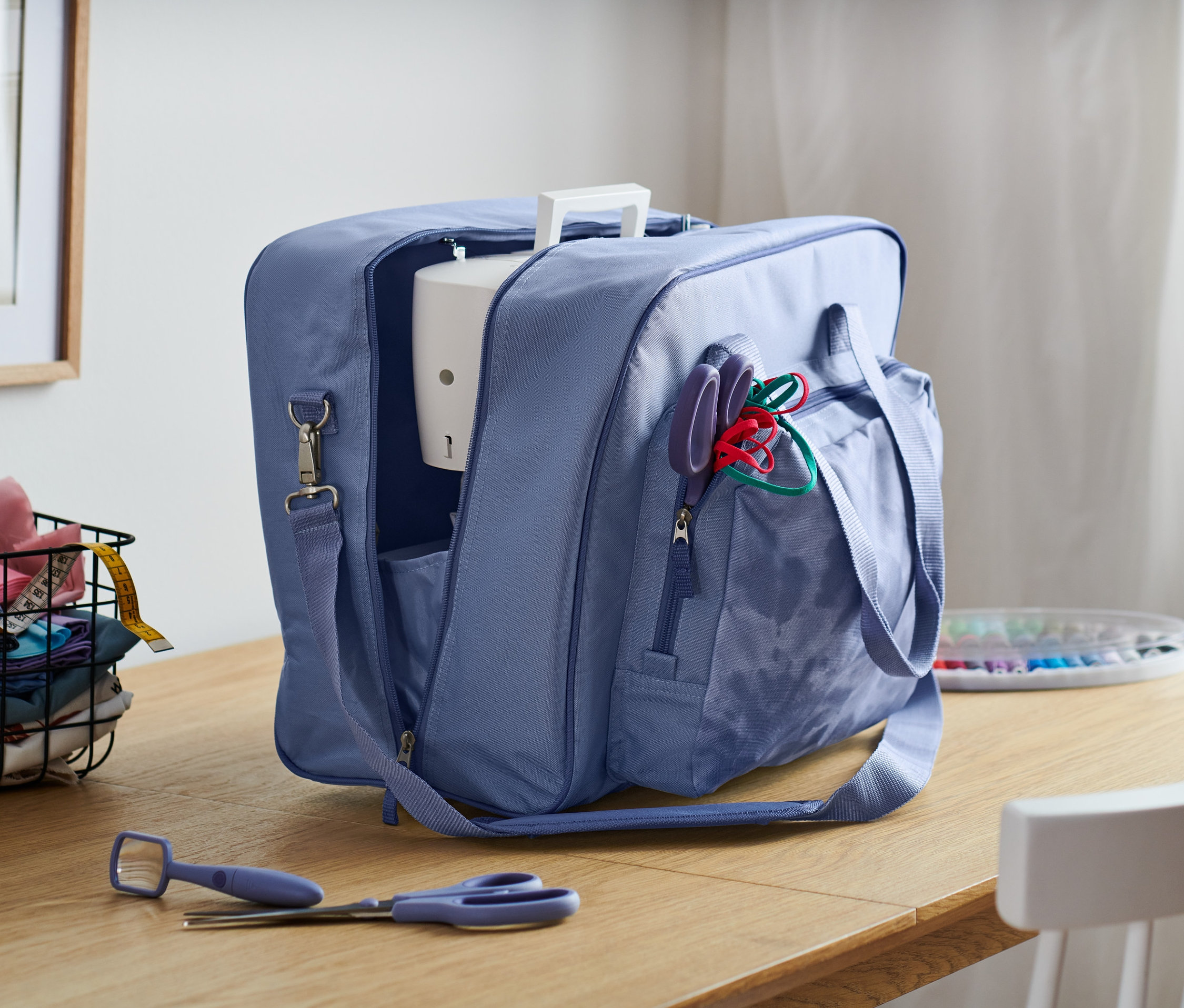 Mavi dikiş makinesi çantası ahşap bir masa üzerinde açık, arka planda kumaş sepeti ve pencere.