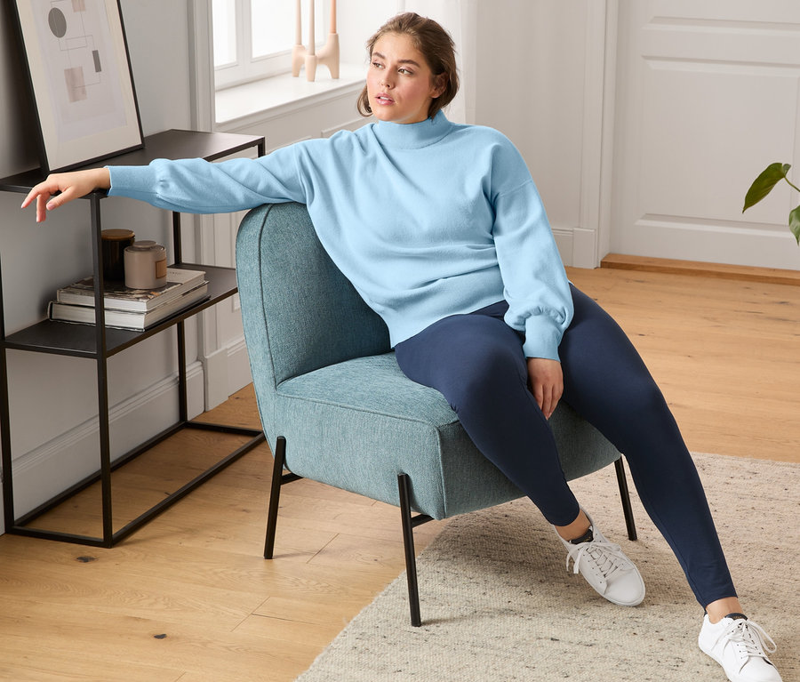 Bir kadın koltukta oturuyor ve kromsuz tabaklanmış deriden yapılmış sneaker, tayt ve balon kollu kazak giyiyor.