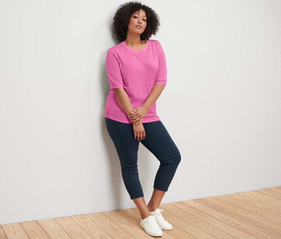 Duvara yaslanmış bir kadın 7/8 Tayt ve İnce Örgülü Kazak giyiyor.