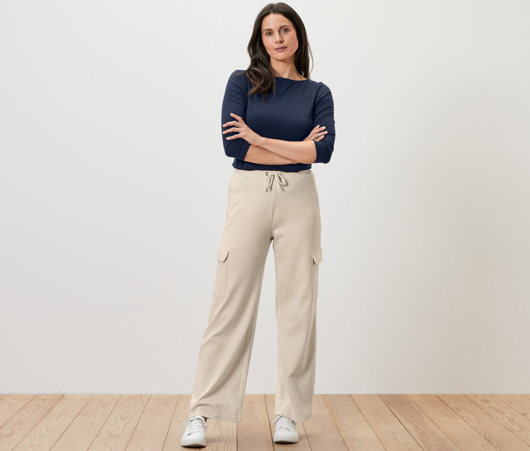 Bir kadın kolları kavuşturmuş şekilde duruyor, üzerinde krem rengi kargo cepli eşofman altı ve lacivert bir üst var.