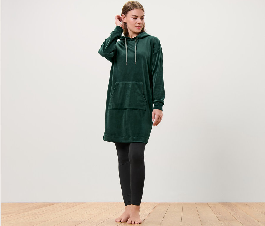 Kadın ayakta duruyor, kapüşonlu yeşil bir Niki Loungewear Elbise ve siyah tayt giyiyor, saçını düzeltiyor.