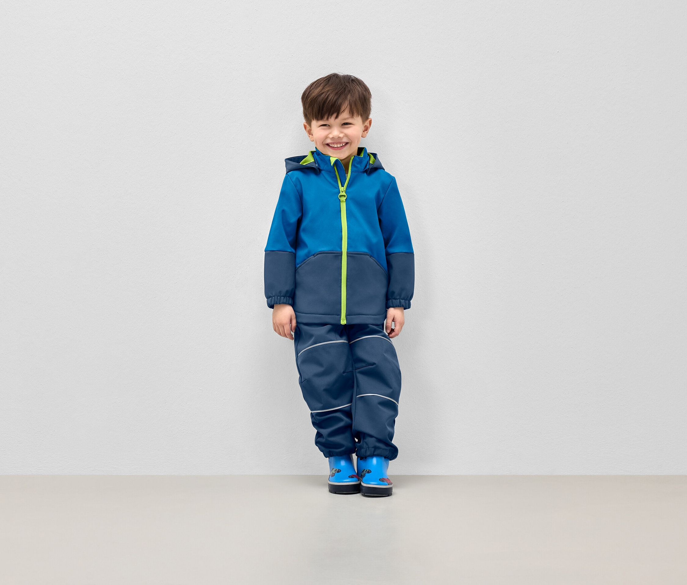 Gülümseyen çocuk, kapüşonlu mavi bir ceket ve reflektörlü şeritleri olan lacivert pantolonla poz veriyor.