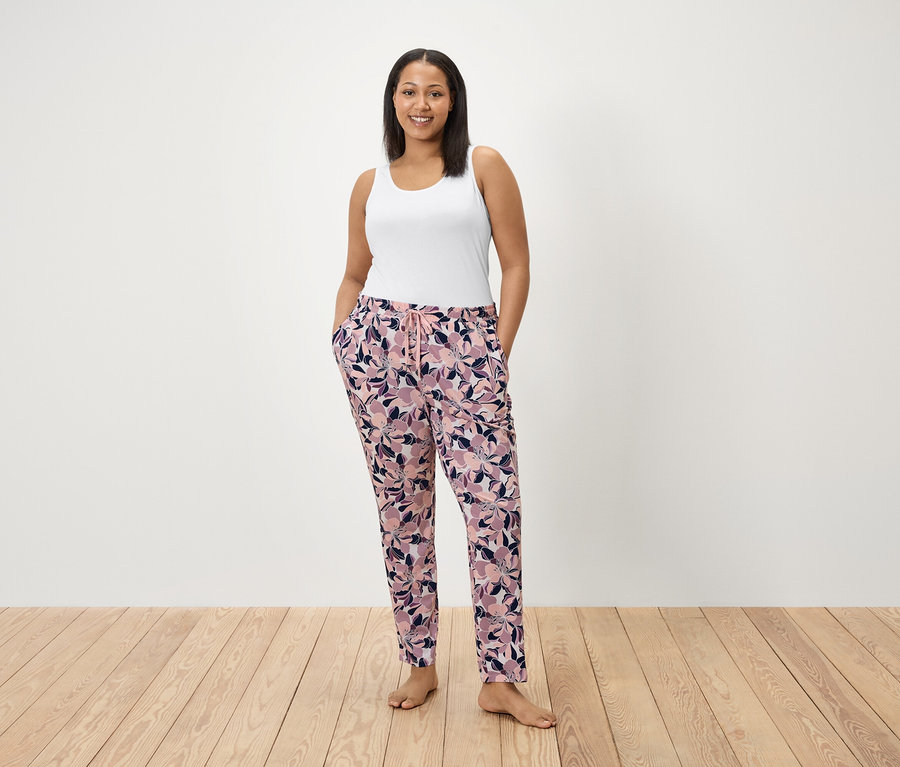Bir kadın çiçek desenli pantolonunun ceplerinde elleriyle ayakta duruyor. Beyaz bir atlet giyiyor.