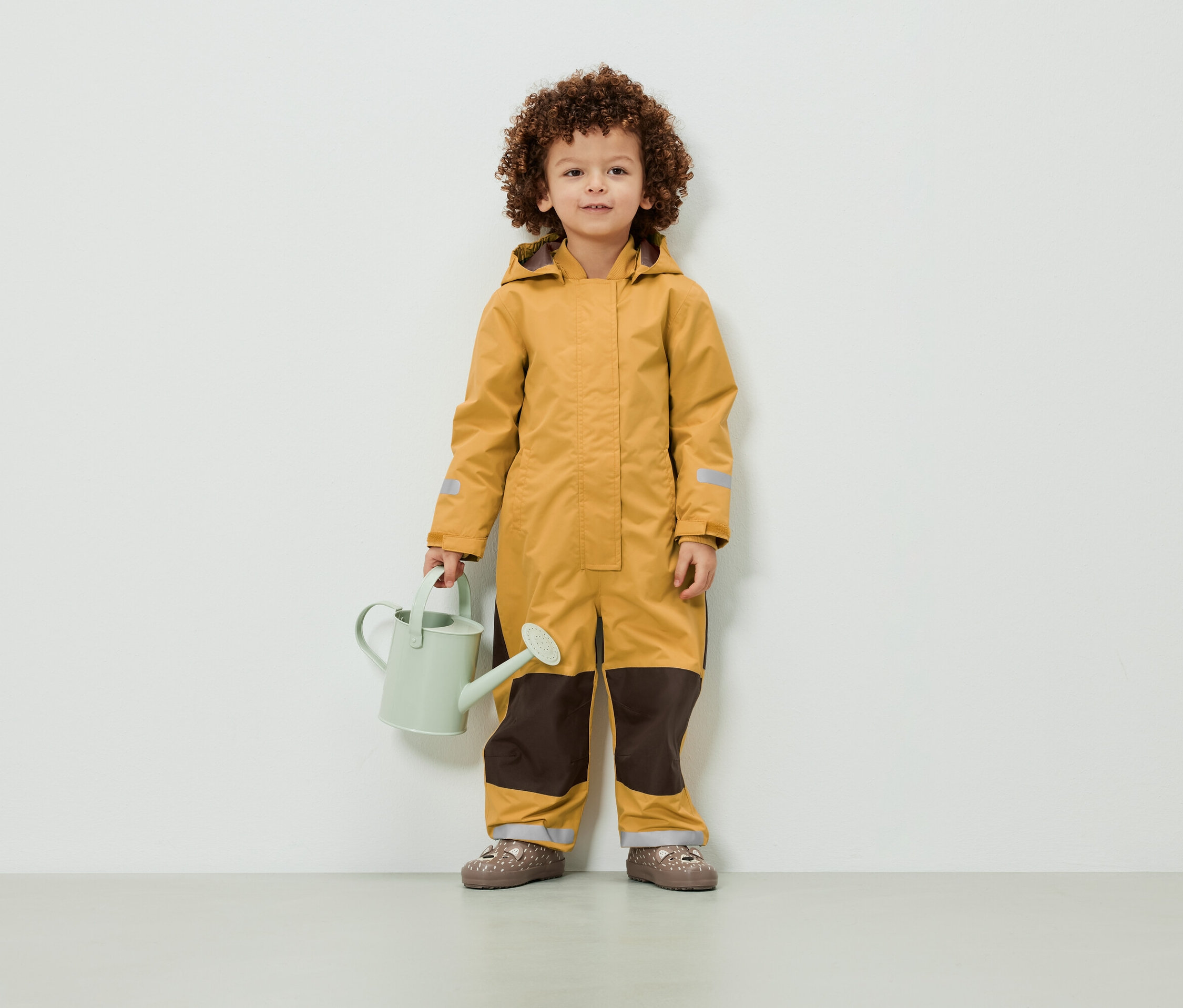 Kıvırcık saçlı bir çocuk sarı bir tulum giymiş ve açık yeşil bir sulama kabı tutuyor.