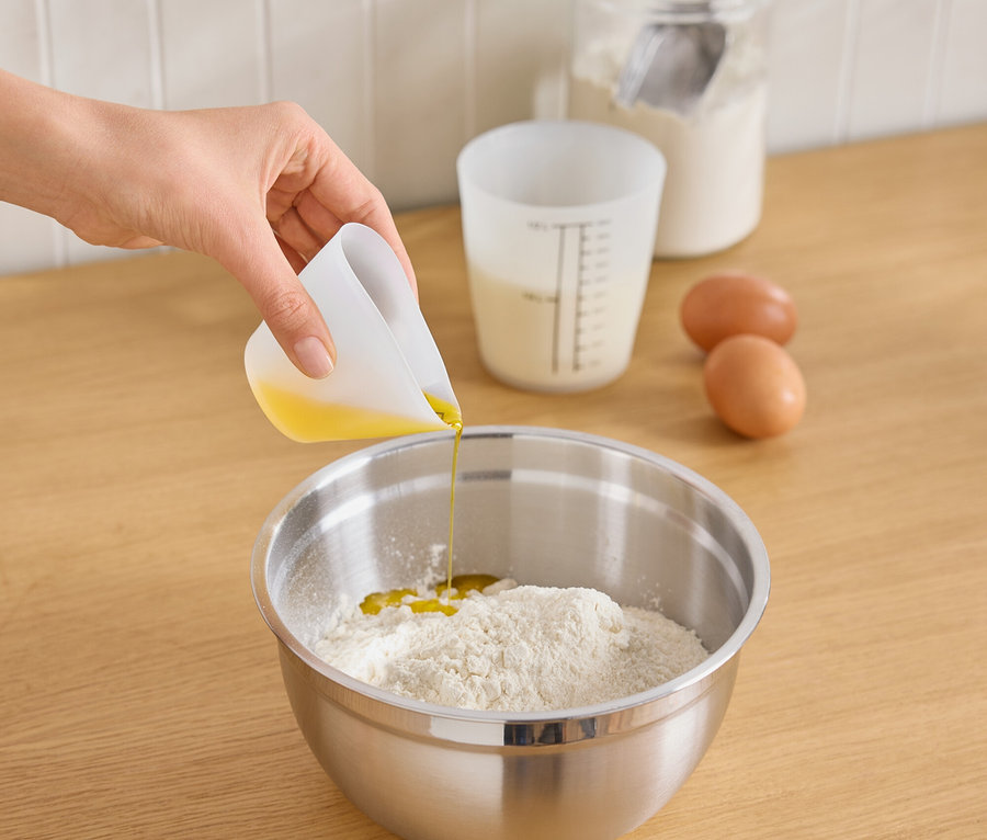 Bir el, beyaz bir ölçüm kabından paslanmaz çelik bir kaseye unun üzerine yağ döküyor. Tezgahta iki yumurta, sütlü bir ölçüm kabı ve un ve kaşıklı bir kavanoz var.