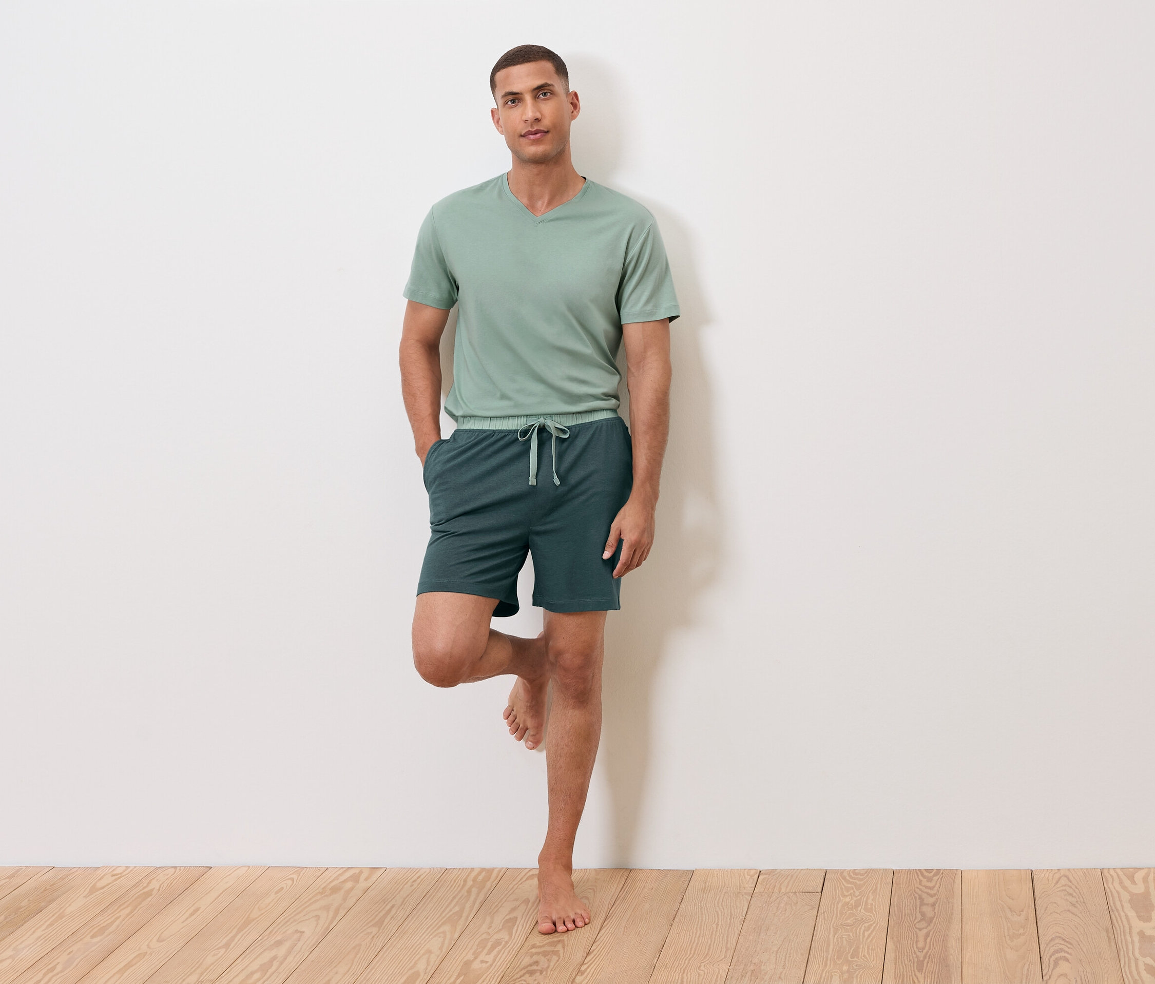 Bir adam bir ayağı duvarda duruyor, üzerinde yeşil bir tişört ve şort var.