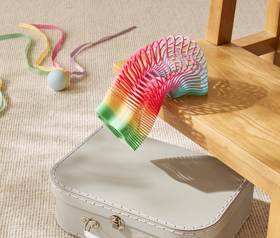 Gökkuşağı renkli oyuncak yay, ahşap bir sandalyenin altında bej bir bavulun üzerinde kısmen duruyor.