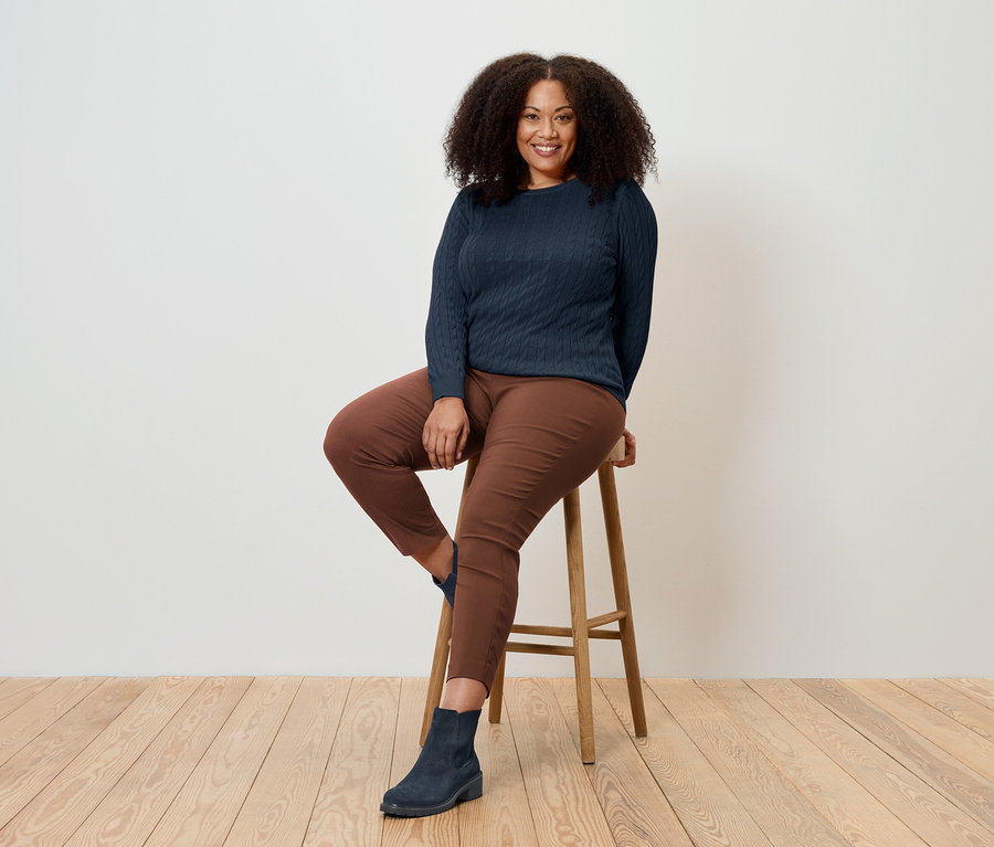 Bir kadın ahşap bir taburede oturuyor, lacivert bir kazak, kahverengi pantolon ve mavi botlar giyiyor.