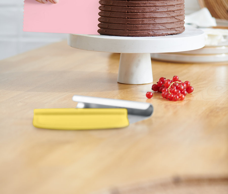 Bir el, pembe bir spatula ile çikolatalı pastanın üzerindeki kremayı düzeltiyor. Pasta beyaz bir stand üzerinde duruyor ve kırmızı frenk üzümü ile süslenmiş. Masanın üzerinde sarı bir spatula duruyor.