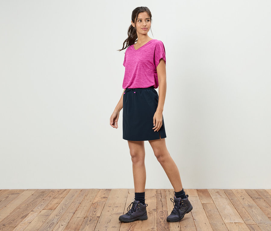 Bir kadın beyaz bir duvarın önünde duruyor, pembe bir tişört, siyah bir etek ve yürüyüş ayakkabıları giyiyor.