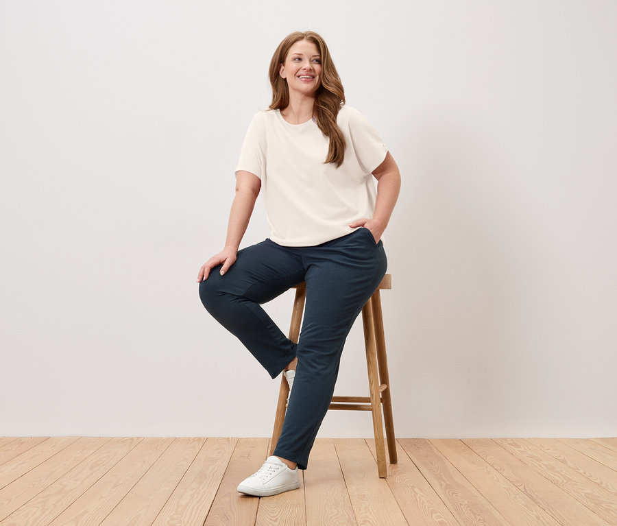 Kadın taburede oturuyor, krem rengi tişört, mavi pantolon ve beyaz spor ayakkabı giyiyor.