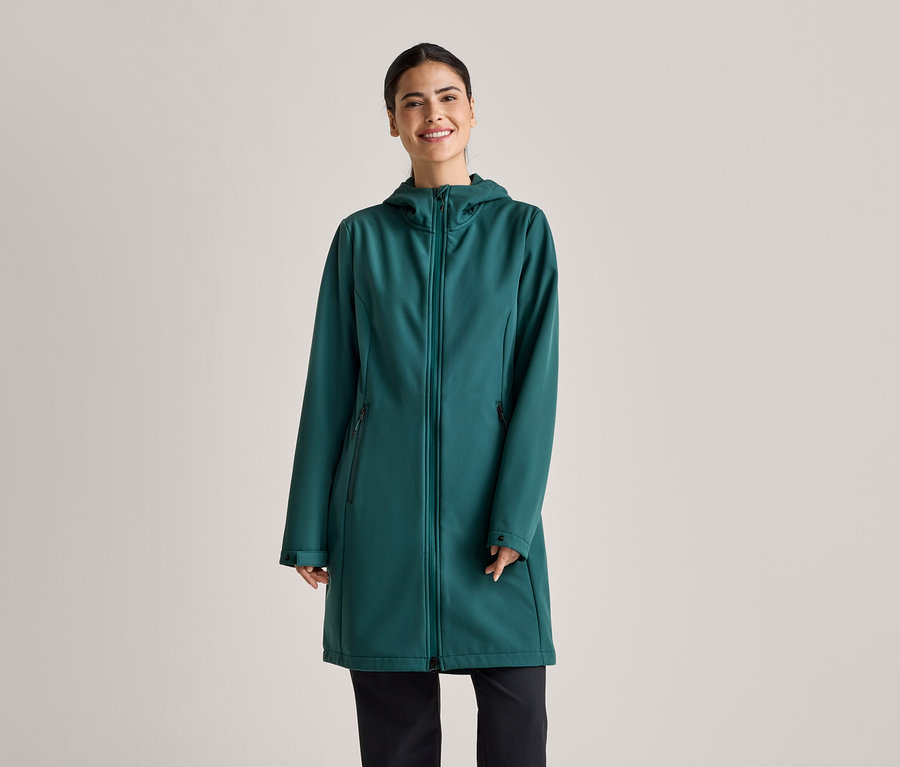 Atkuyruklu kadın, açık gri bir arka plan önünde zümrüt yeşili, kapüşonlu, uzun softshell manto ve siyah pantolon giyiyor.