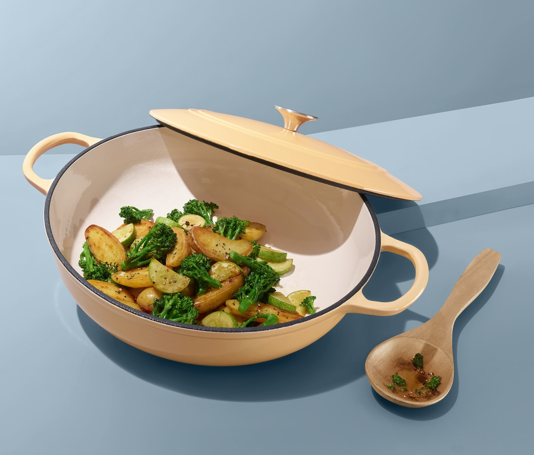 Kapaklı ve içinde pişmiş sebzeler olan sarı kızartma tavası, mavi bir arka plan üzerinde tahta bir kaşığın yanında duruyor.