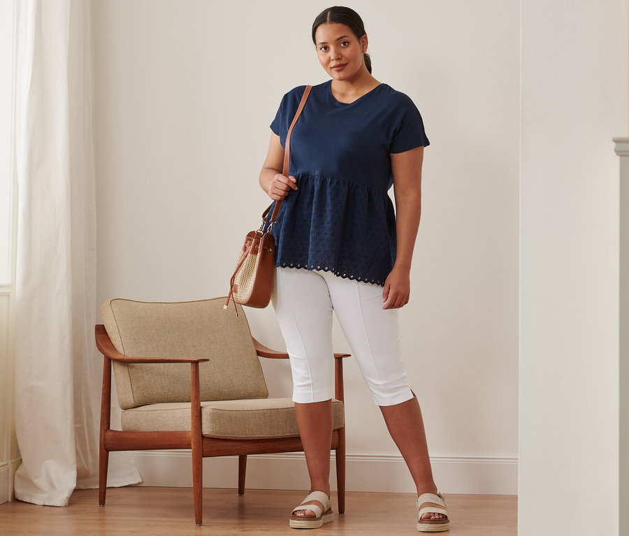 Bir kadın, lacivert renkli, işlemeli, karışık malzemeden yapılmış bir tişört, 3/4 streç pantolon ve büzgülü çanta giyiyor ve ayakta duruyor.