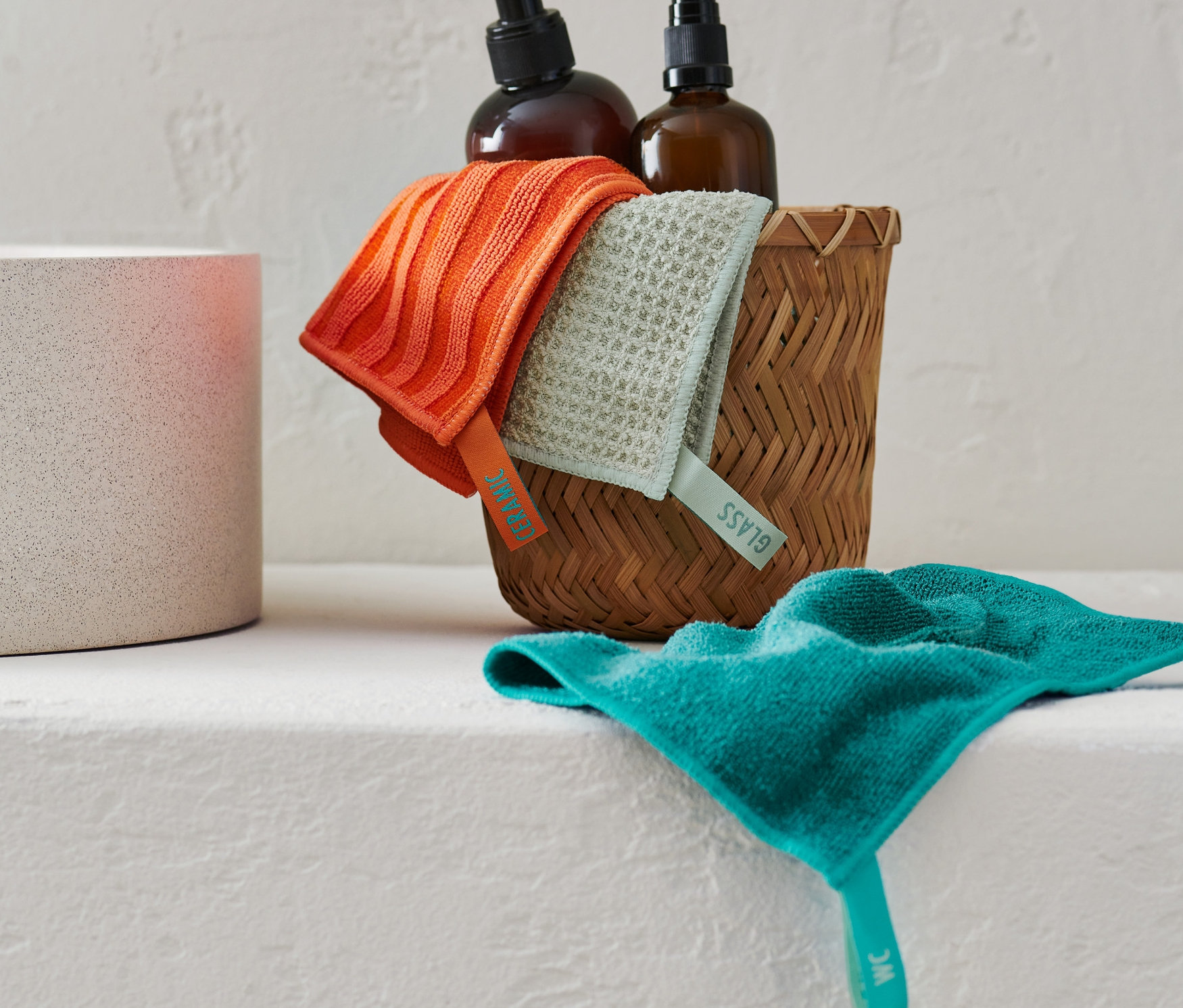 Temizlik bezi seti, iki kahverengi şişenin yanında hasır bir sepetin içinde duruyor.