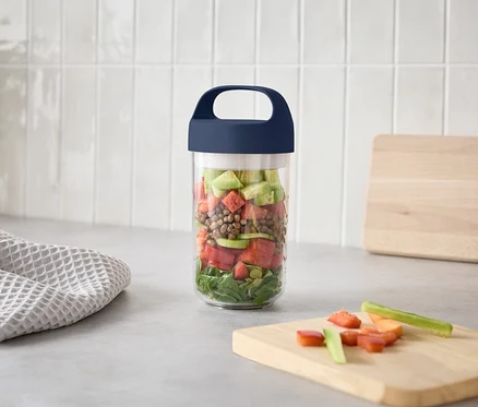Katmanlı salata bulunan bir kavanoz, kesilmiş sebzelerle birlikte bir kesme tahtasının yanında mutfak tezgahında duruyor.