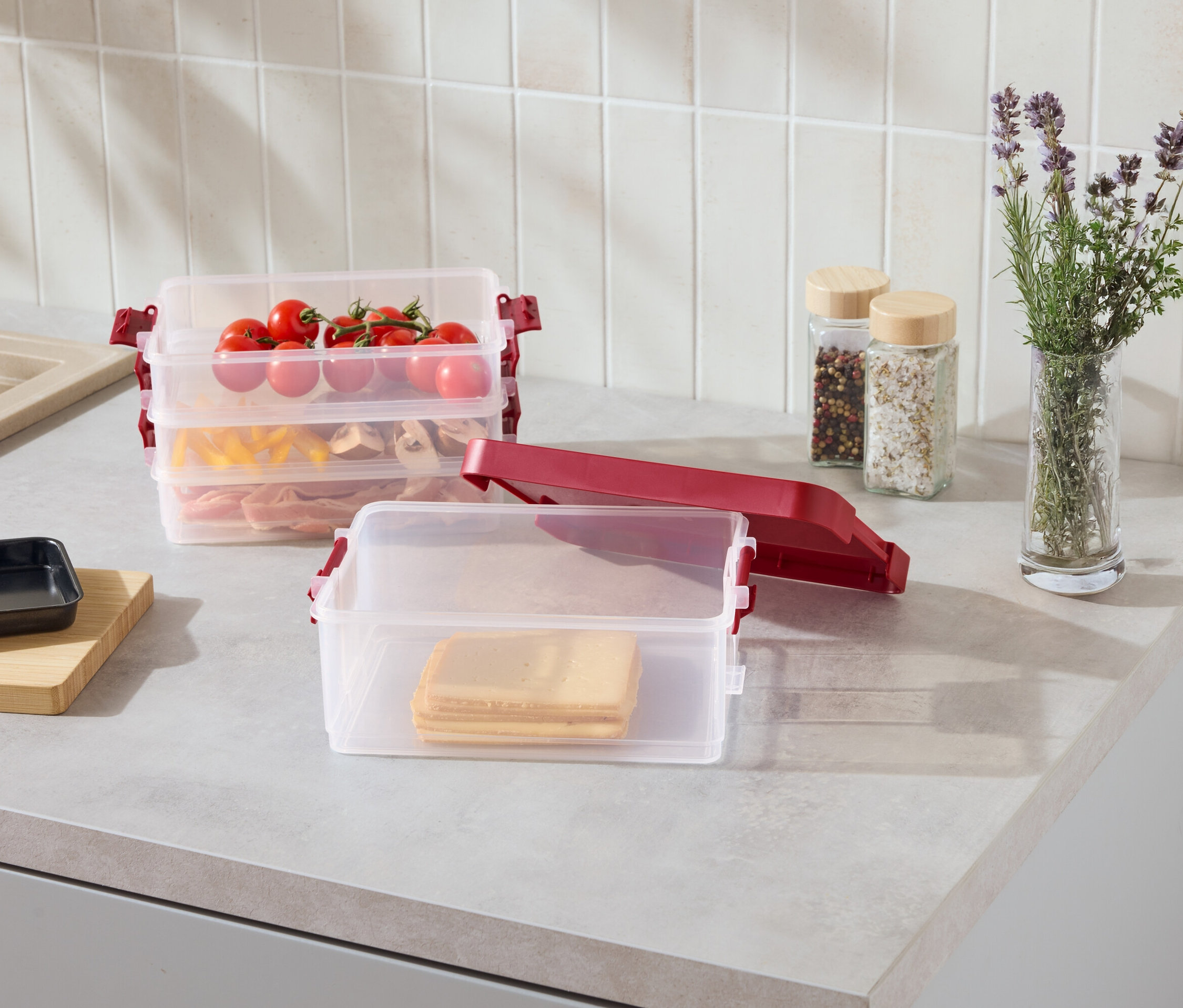 Mutfak tezgahında kırmızı klipsli, şeffaf, istiflenebilir kaplar; içlerinde peynir dilimleri, makarna, et ve domates var.