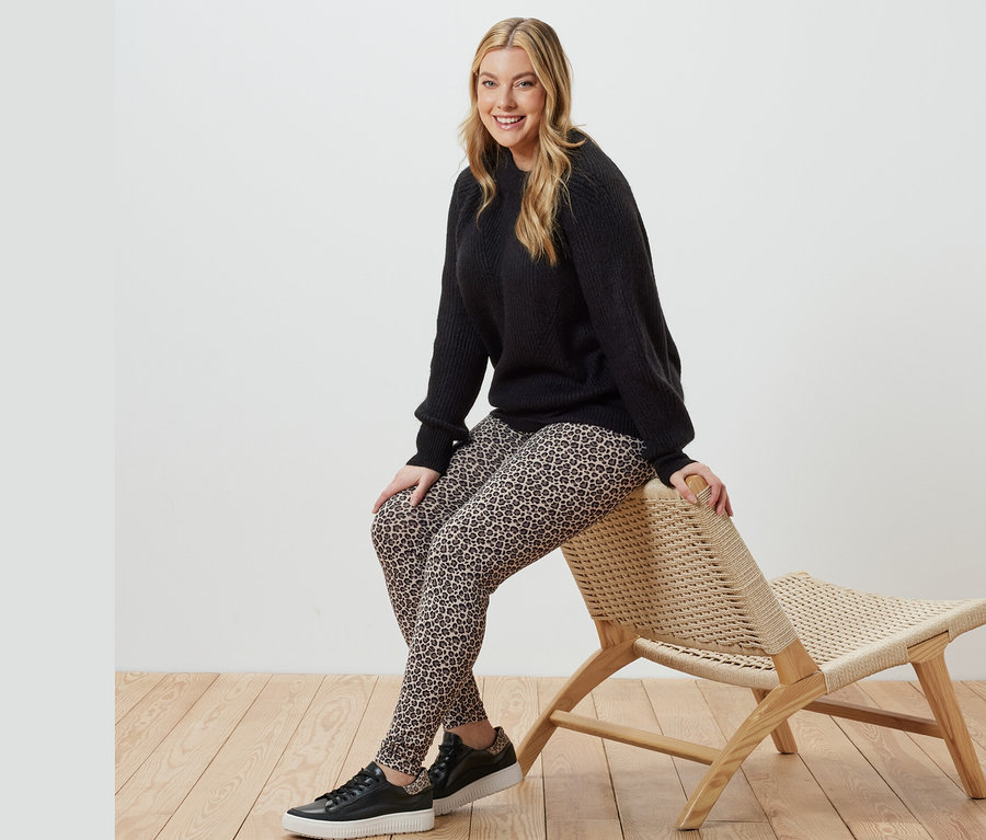 Bir kadın hasır bir sandalyede oturuyor, üzerinde siyah örgü kazak ve desenli tayt var.