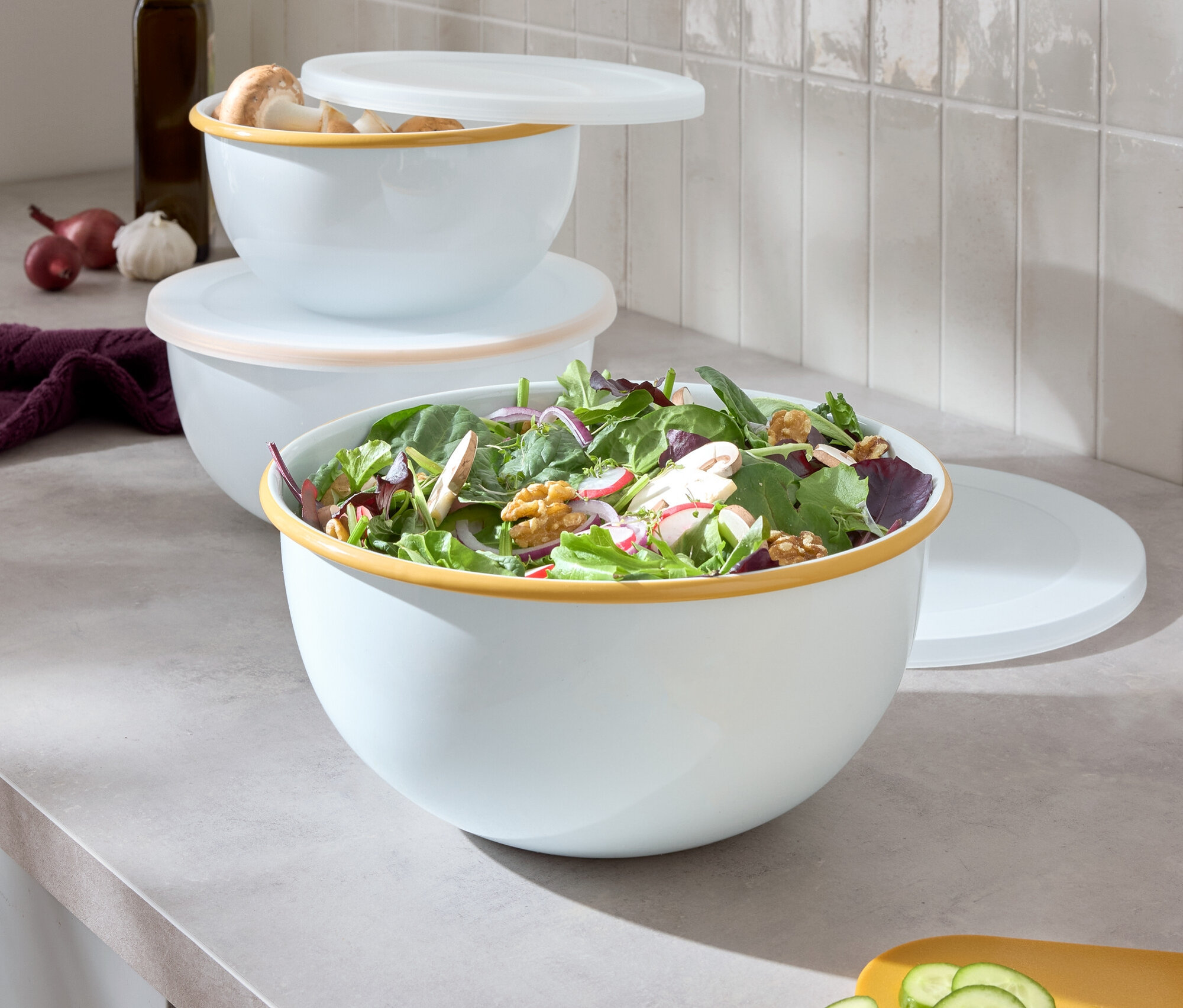 Renkli kenarlı üç beyaz kase üst üste yığılmış, en büyük kase salata içeriyor, yanında kesilmiş salatalık bulunan sarı bir kesme tahtası var.