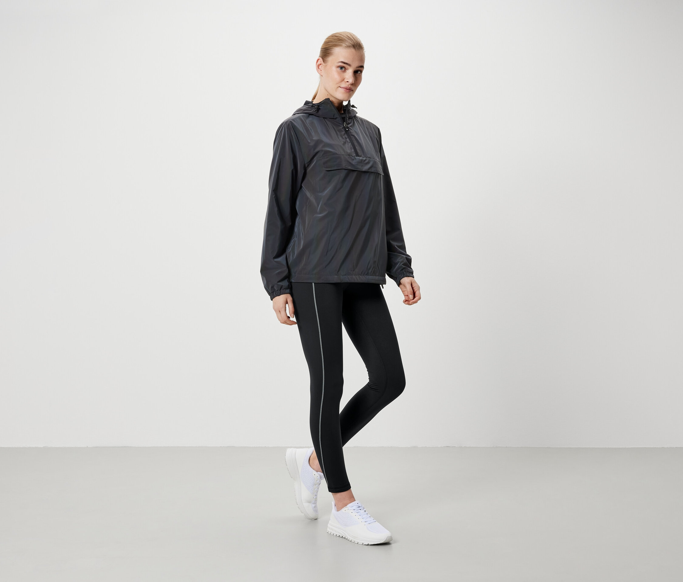 Sarışın bir kadın gri kapüşonlu ceket, siyah tayt ve beyaz ayakkabılarla ayakta duruyor.