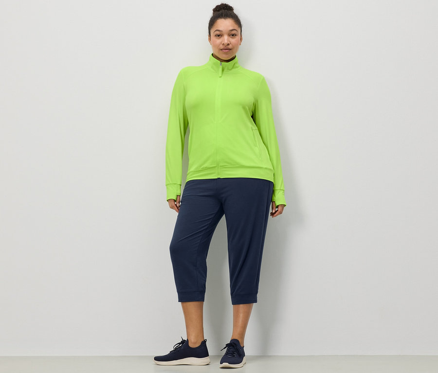 Beyaz bir duvarın önünde duran bir kadın, Limon Yeşili Fonksiyonel Ceket ve Lacivert 3/4 Sweat Pantolon giyiyor.