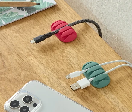 Bir masa üzerinde pembe ve yeşil renklerde iki adet Universal-Kabelmanager, bir telefon ve kalemler bulunmaktadır.