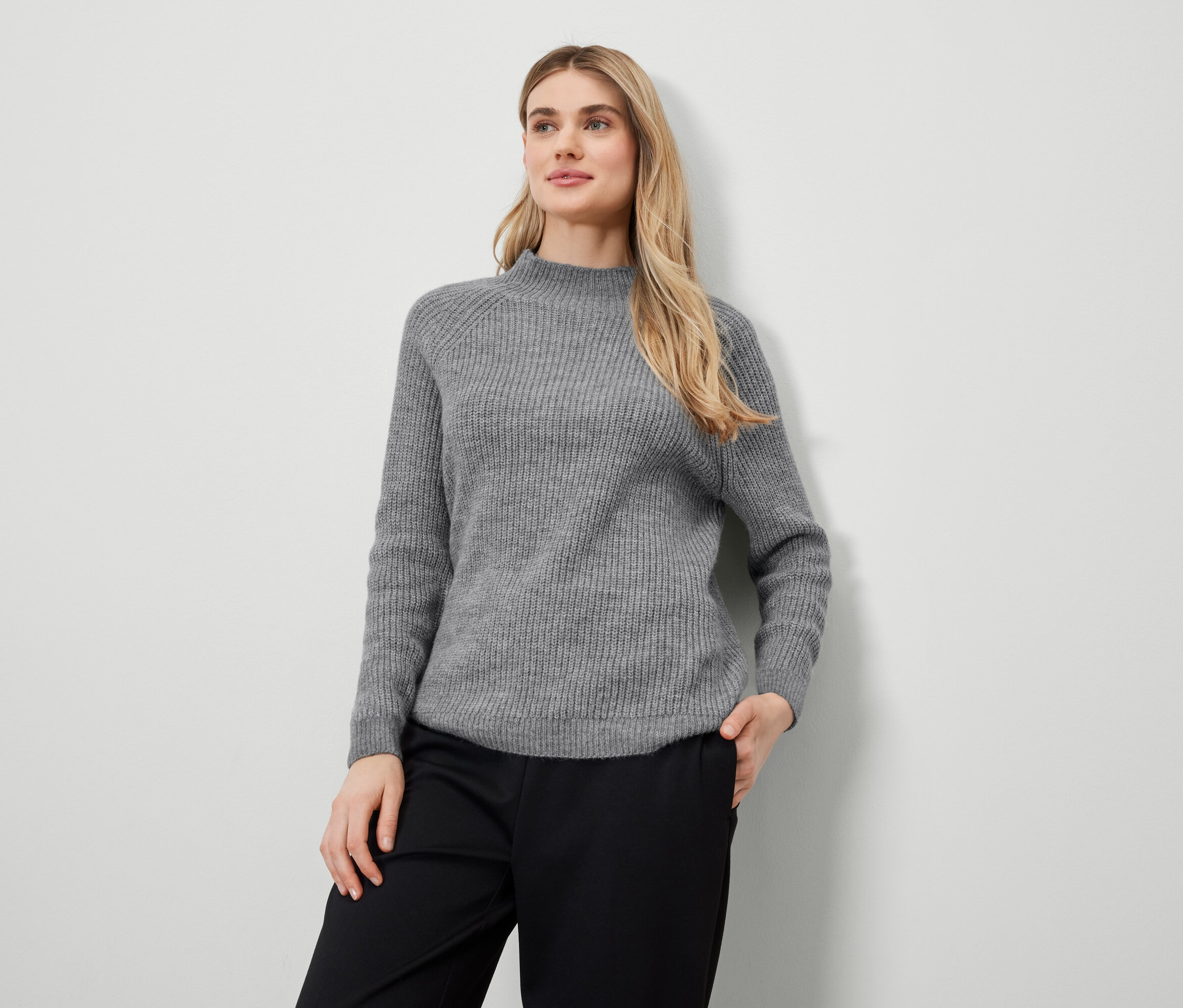 Sarışın kadın gri kazak ve siyah pantolonla poz veriyor.