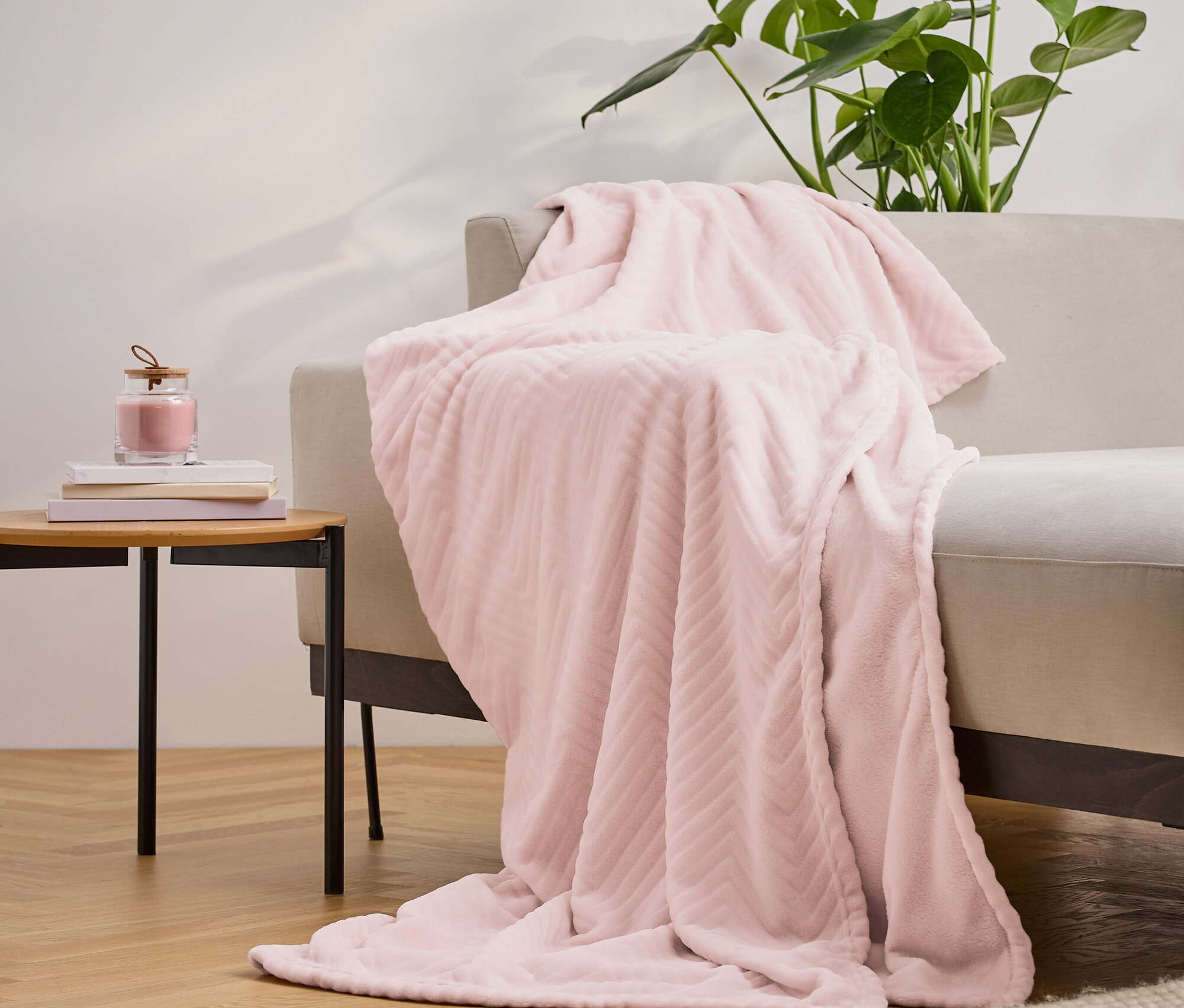 Bej bir kanepenin üzerine atılmış pembe, dokulu bir battaniye, yanında kitapların ve bir mumun bulunduğu bir masa ve arka planda bir bitki. 