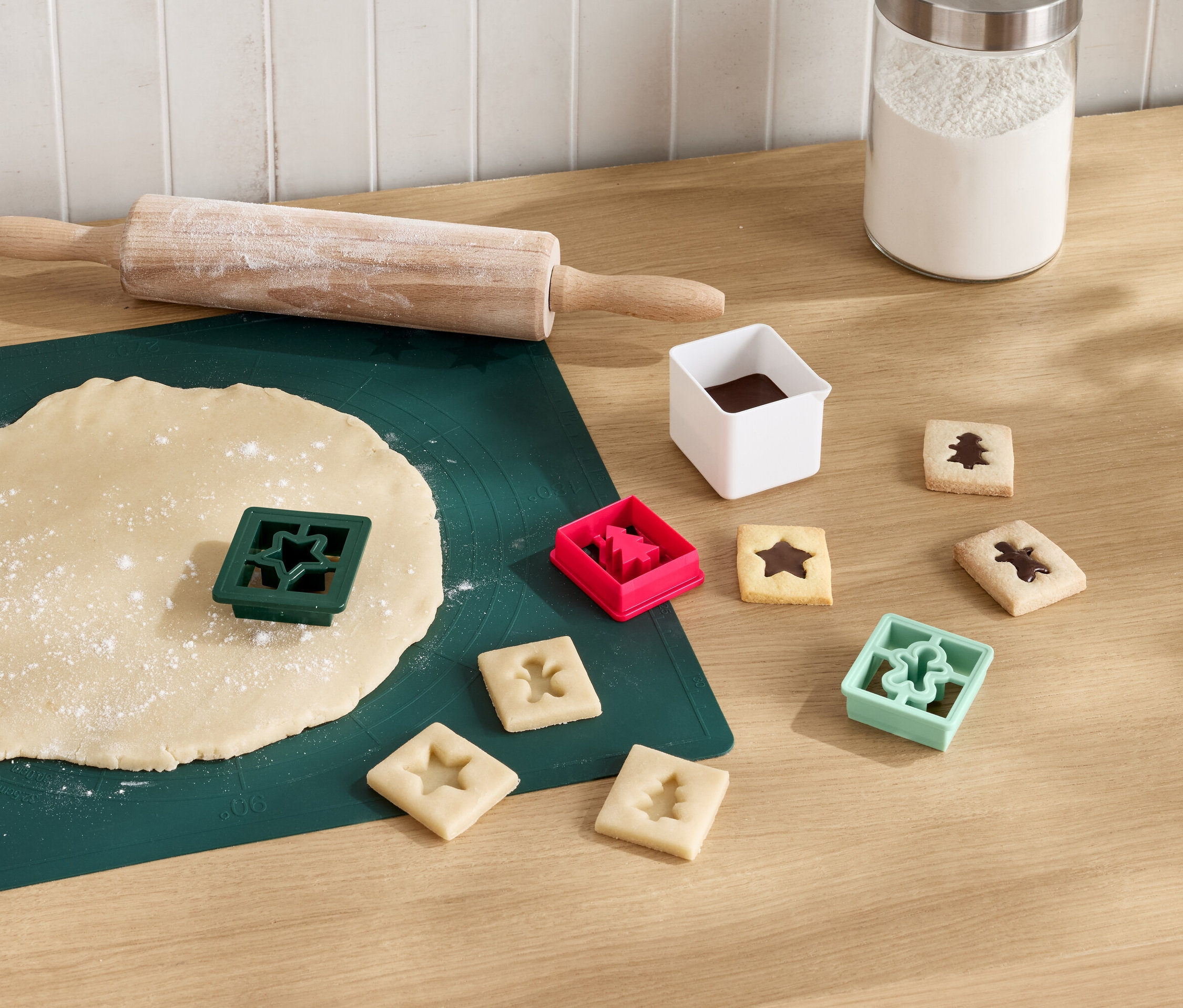 Yeşil bir mat üzerinde açılmış hamur, oklava ve yıldız şeklinde kurabiye kalıbı. Yanında çikolatalı kesilmiş kurabiyeler ve ağaç, yıldız ve insan figürlü kalıplar bulunuyor.