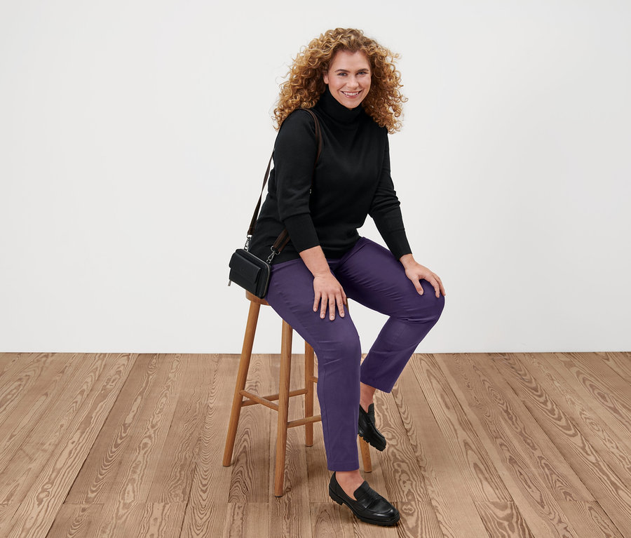 Kadın bir taburede oturuyor, üzerinde siyah ince triko balıkçı yaka kazak, mor pantolon ve siyah ayakkabılar var.