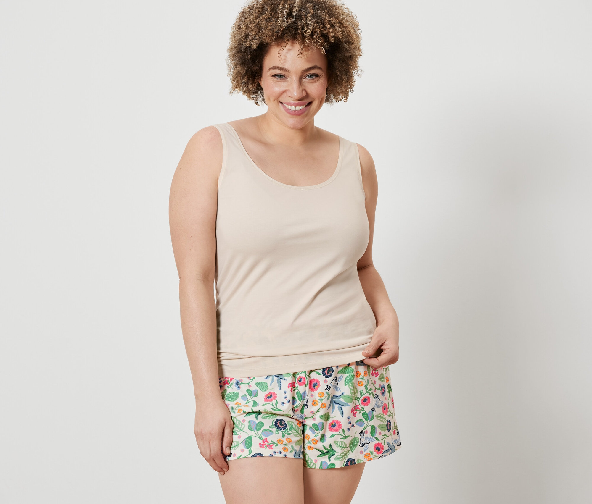 Kadın, 2 Adet Şort ve krem rengi bir atletten oluşan çiçekli şort takımı giyiyor.