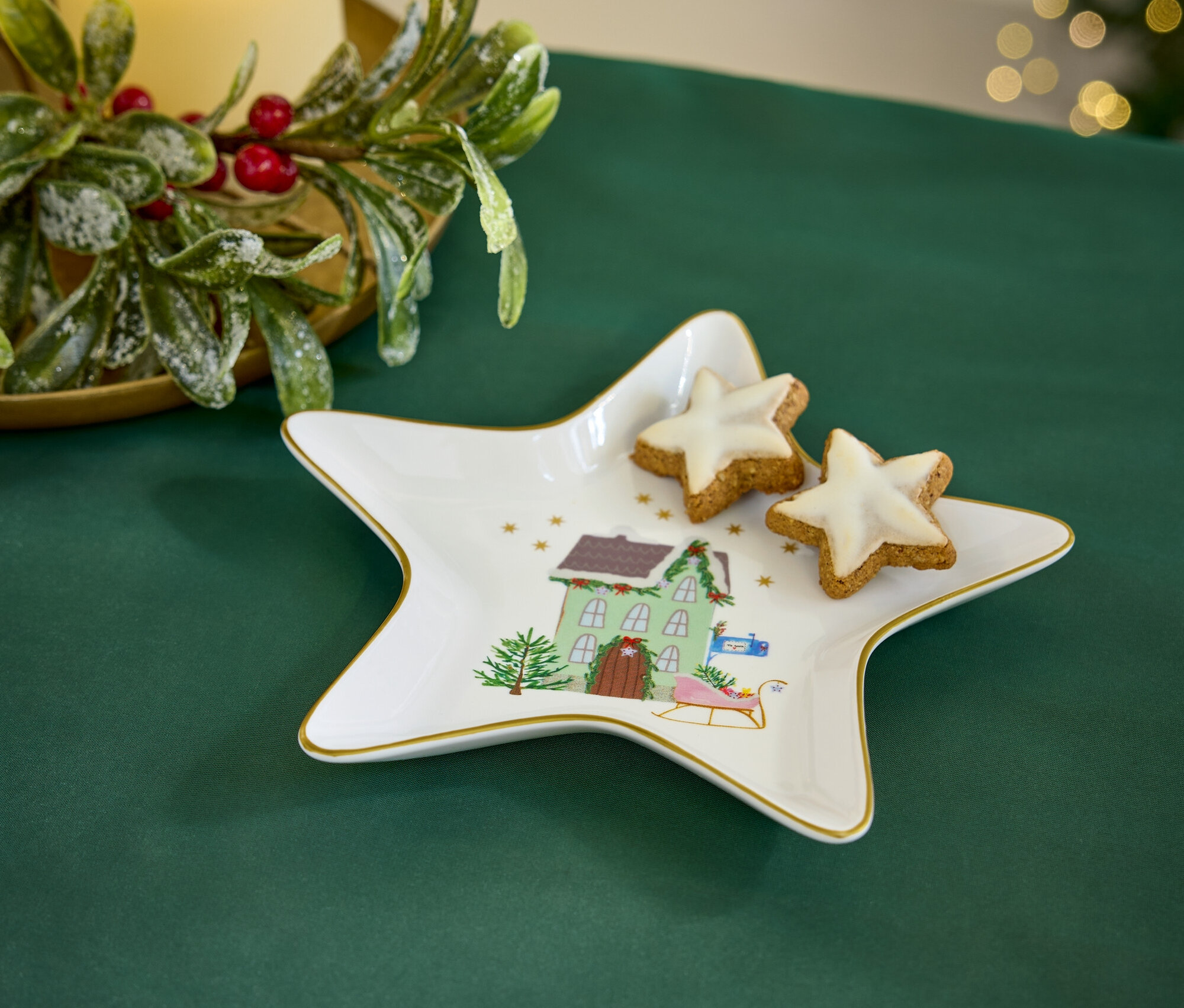 Yeşil bir örtü üzerinde, Noel temalı yıldız şeklinde bir tabak ve iki yıldız şeklinde kurabiye bulunmaktadır. Arka planda bir mum ve Noel dekorasyonu görülmektedir.