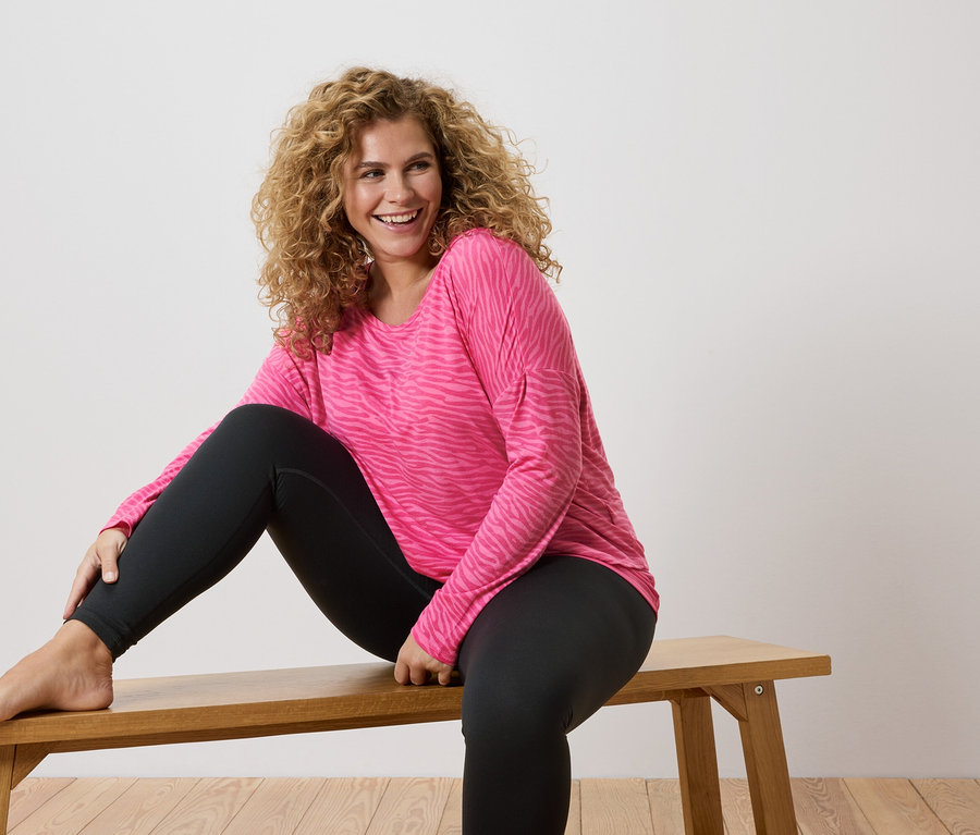 Kıvırcık saçlı kadın, pembe zebra desenli uzun kollu bir tişört ve siyah tayt giyerek çıplak ayakla ahşap bir bankta oturuyor.