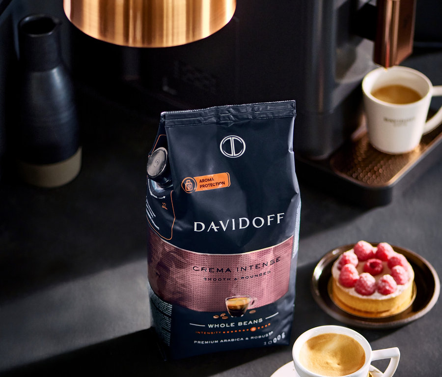Bir paket DAVIDOFF CAFÉ Crema Intense 1000 g bütün çekirdek kahve, bir fincan kahve ve bir ahududulu tartın yanında duruyor.