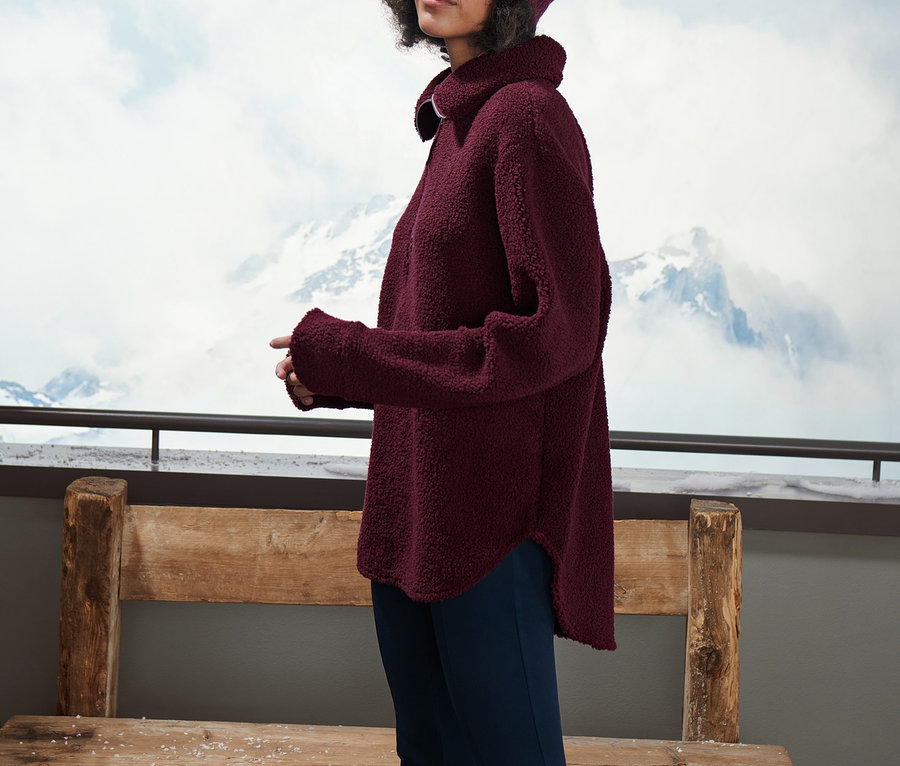 Dağ manzarası önünde bordo büyük beden pelüş polar ve uyumlu bere giyen bir kadın poz veriyor.