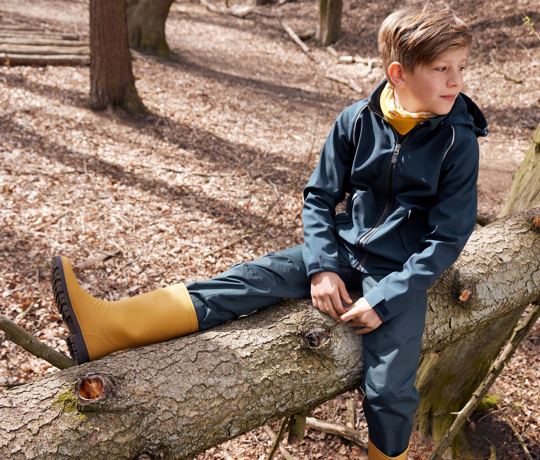 Ormanda bir ağaç kütüğünde oturan bir çocuk. Üzerinde Softshell ceket, yağmur pantolonu, sarı geri dönüştürülmüş lastik çizme ve çok fonksiyonlu boyunluk var.