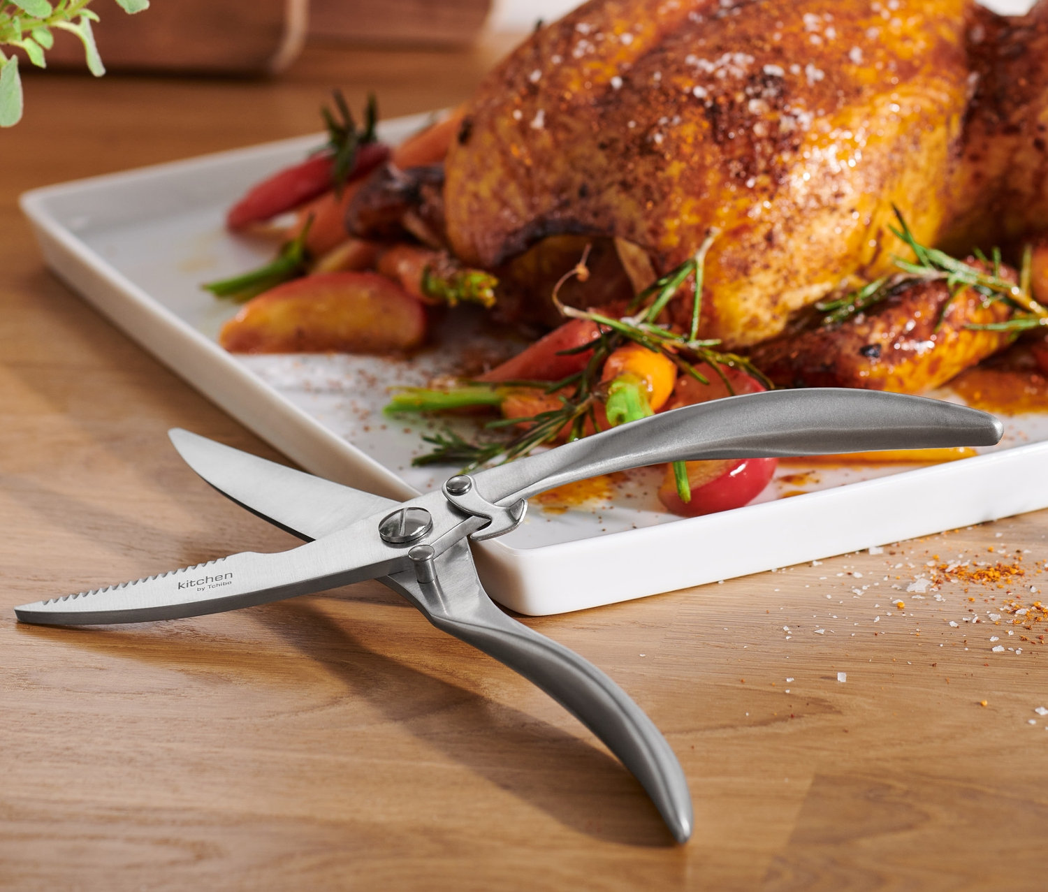 Ahşap bir masanın üzerinde havuç ve otlar ile birlikte beyaz bir tabakta kızarmış bir tavuk ve bir Tavuk Makası bulunmaktadır.