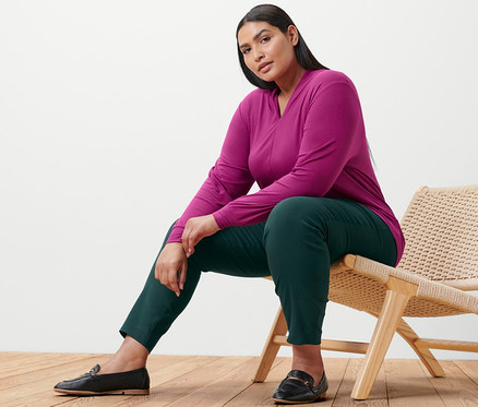 Kadın sandalyede oturuyor. Üzerinde böğürtlen rengi, V yakalı uzun kollu bir tişört ve streç pantolon var.